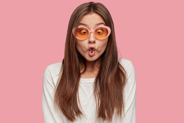 Funny playful Caucasian woman makes fish lips, has crazy look, tries to amuse depressed friend, feels bored, wears trendy shades and white sweater, isolated over pink background. Facial expressions - obrazy, fototapety, plakaty