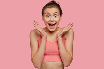 Isolated shot of overjoyed smiling young female fitness model in pink top clasps hands, has sporty...