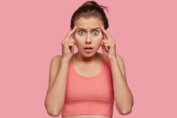 Photo of stressed overworked young female athlet keeps both index fingers on temples, has headache,...