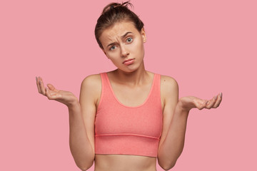 Indifferent Caucasian woman shrugs shoulders with hesitation, cant make choice which sport to choose, dressed in casual top, isolated over pink background. Athletic girl expresses uncertainty