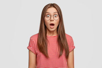 Studio shot of stupefied emotive woman keeps jaw dropped, reacts on sudden news from friend, wears...