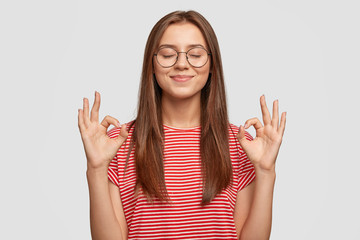 Pleased satisfied young female model makes zero gesture, wears transparent glasses, has long dark...