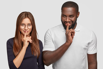 Mysterious multiethnic woman and man look secretly at camera, keep both index fingers on lips as try be mute, tell private information, model against white background. Keep voice down, please