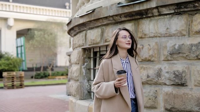Attractive European brunette girl with glasses, brown coat and stylish look. A business lady tapping the message on her phone, and has a coffee break of the sunset and city center. autumn style