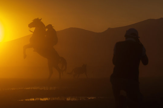 Silhouette of a cowboy puting his horse to stay in two feets at sunset with dust in background and a photographer taking pictures of him