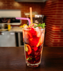 a glass of beautiful bright cocktail on a wooden surface decorated with fruits, berries, leaf of mint and basil at bar counter background