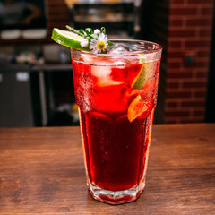 a glass of beautiful bright cocktail on a wooden surface decorated with fruits, berries, leaf of mint and basil at bar counter background