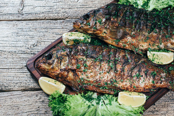 A deliciously roasted carp on a grill, presented on a wooden board, and along the leaves of green salad and pieces of lemon