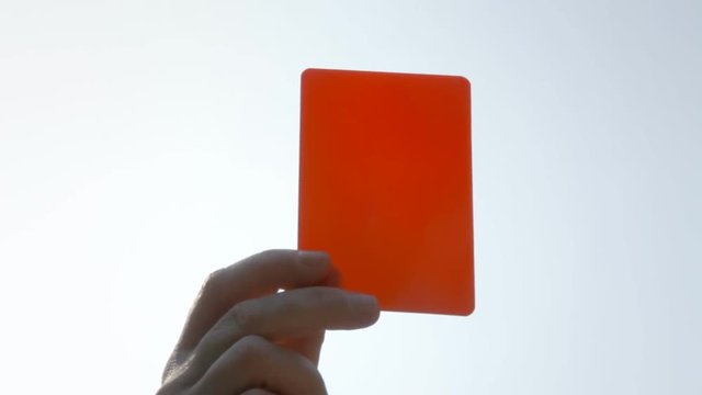 The hand of a referee showing a red card against a clean sky, during a soccer or football match. Close-up shot.
