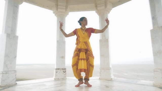 A wide shot of a Indian folk dancer dancing elegantly in a white marble temple located on a hilltop. A movement shot of a beautiful woman in traditional yellow sari performing classical dance form