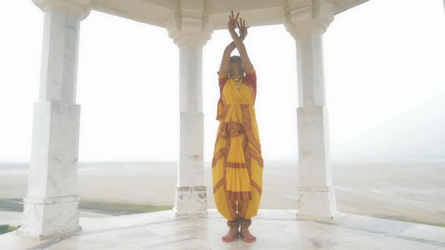 A movement shot of a beautiful woman in traditional yellow sari performing classical dance form. A wide shot of a Indian folk dancer dancing elegantly in a white marble temple located on a hilltop.