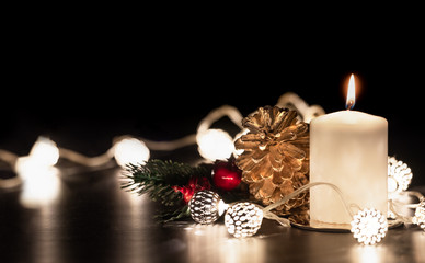 Christmas white candle with golden pine cone,mistletoe and light string glowing on black marble table with dark background.holiday greeting card.