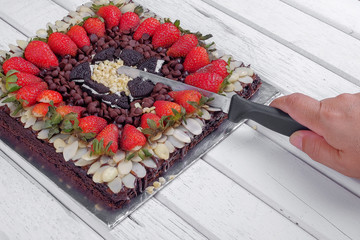 A man's hand with a kitchen knife above a brownie cake with strawberries on a wooden table