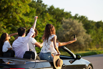 Company of young women and guys are making selfie in a black cabriolet on the road on a warm sunny...