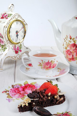 Piece of delicious homemade brownie cake with strawberries , vintage table clock and tea set on white table. Selective focus