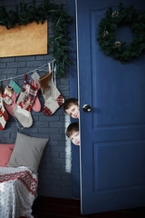 Two boys peeking out from behind the open door in the gray room of the Christmas decoration.