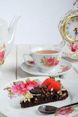 Piece of delicious homemade brownie cake with strawberries , vintage table clock and tea set on white table. Selective focus