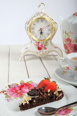 Piece of delicious homemade brownie cake with strawberries , vintage table clock and tea set on white table. Selective focus
