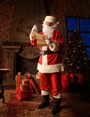 Santa Claus standing at his room at home near Christmas tree and big sack and reading Christmas...