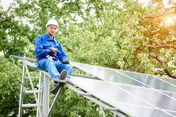 Young smiling electrician points screwdriver at camera sitting on almost finished stand-alone solar photo voltaic panel system on bright sunny green tree background. Alternative energy concept.