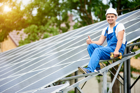 Young smiling electrician sitting on almost finished stand-alone solar photo voltaic panel system with screwdriver, showing thumb up gesture on bright sunny green tree background