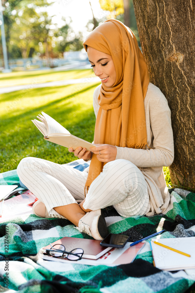 Wall mural Photo of young arabic female student wearing headscarf, sitting on blanket in green park and reading book