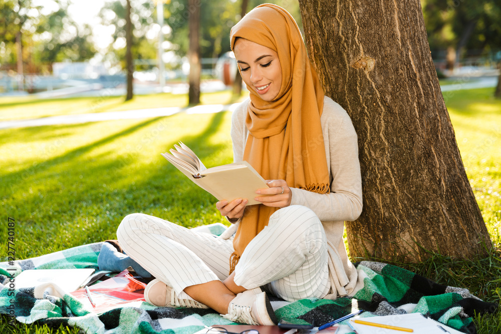 Wall mural Photo of muslim girl student wearing headscarf, sitting on blanket in green park and reading book