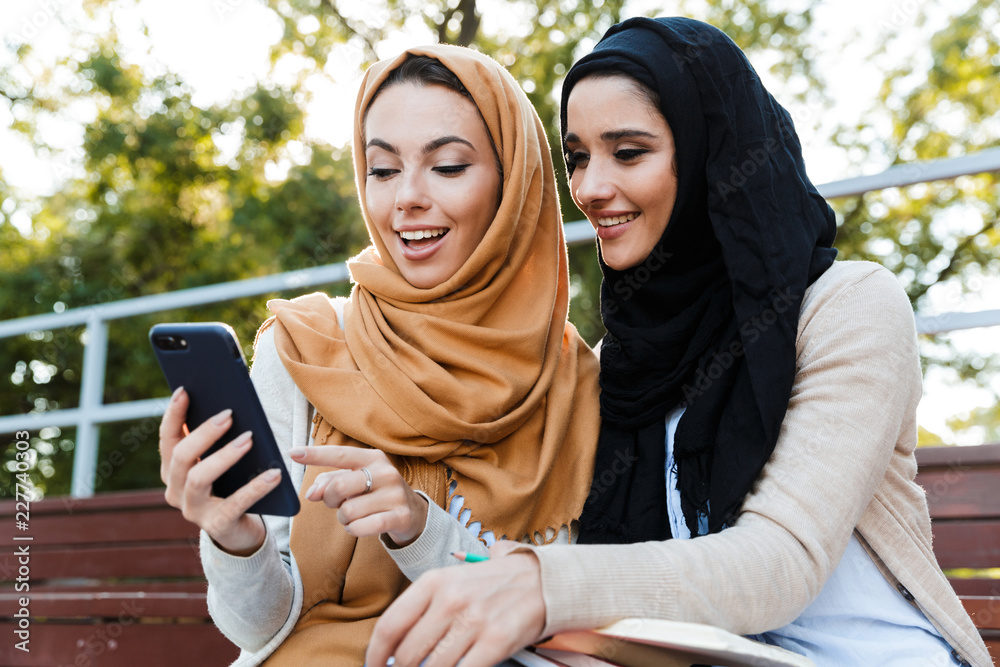 Wall mural photo of beautiful muslim girls wearing headscarfs, sitting in green park and using cell phone