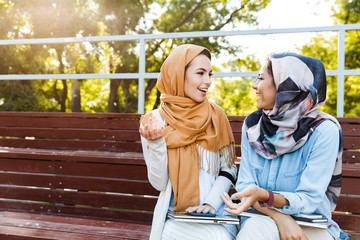 Photo of young islamic girls wearing headscarfs, sitting on bench in park and talking or discussing