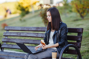 Caucasian woman sitting in park with laptop and eating croissant
