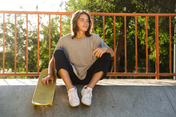Photo of european skater boy 16-18 in casual wear sitting on ramp with skateboard in skate park, during sunny summer day - obrazy, fototapety, plakaty