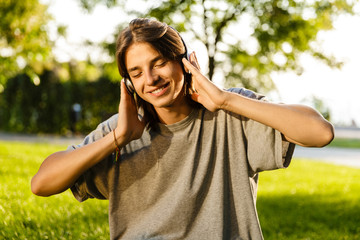 Photo of happy boy 16-18 in casual wear sitting on grass in green park, and listening to music using earphones