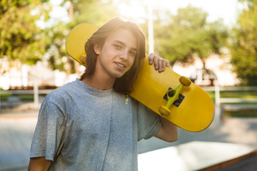 Photo of beautiful skater boy 16-18 in casual wear standing with skateboard in skate park, during sunny summer day © Drobot Dean