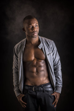 Handsome shirtless muscular black young man's with jacket open on naked torso, looking at camera, on dark background in studio shot