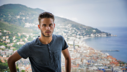 Fototapeta na wymiar Handsome Young Man in Trendy Attire, in a Sunny Summer Day with Italian Sea Coast in the Distance, Wearing a White Shirt
