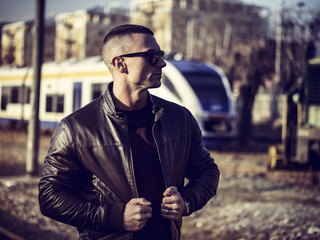 One handsome young man in urban setting in European city in front of train, standing, wearing black leather jacket