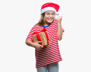 Young beautiful girl wearing christmas hat and holding gift over isolated background screaming proud and celebrating victory and success very excited, cheering emotion