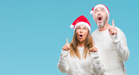 Middle age hispanic couple wearing christmas hat over isolated background amazed and surprised looking up and pointing with fingers and raised arms.