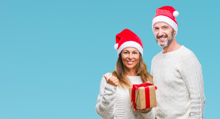 Middle age hispanic couple wearing christmas hat and holding gift over isolated background pointing and showing with thumb up to the side with happy face smiling