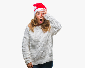Middle age senior hispanic woman wearing christmas hat over isolated background doing ok gesture shocked with surprised face, eye looking through fingers. Unbelieving expression.