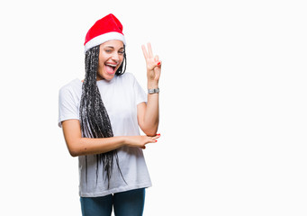 Young braided hair african american girl wearing christmas hat over isolated background smiling with happy face winking at the camera doing victory sign. Number two.