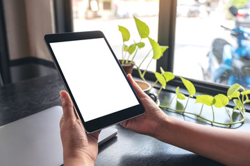 Mockup image of hands holding black tablet pc with blank white desktop screen with green leaves on wooden table