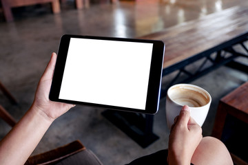 Mockup image of woman's hands holding black tablet pc with blank white screen while drinking coffee on wooden table in cafe