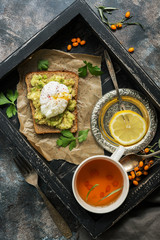Breakfast poached egg, avocado guacamole sandwich, tea, lemon on a wooden tray. Rustic background. Tea with sea buckthorn. Top view, flat lay.