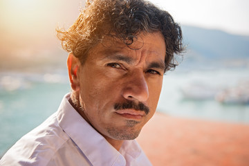 Portrait of a man with an interesting face typical of southern Italy, that can be defined both handsome or ugly, with black hair, moustaches, white skirt, braces, leaning to a wall.