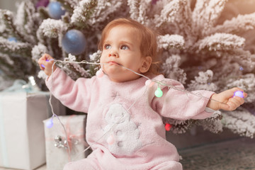 Small girl in the pink pajama sitting near the christmas tree playing with the garland