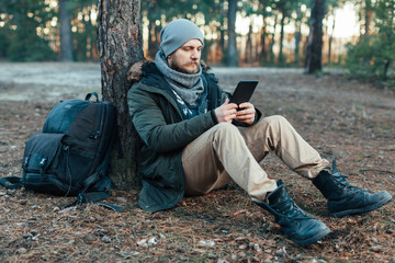 adult handsome Male traveler Sits On autumn pine forest Near tree, Holding At Tablet Recreation...
