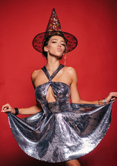 Beautiful young woman in black witch costume with hat and standing over red background. Elegant...