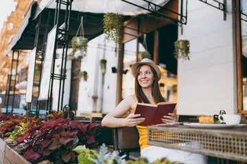 Woman in outdoors street coffee shop cafe sitting at table in hat, reading book with cup of cappuccino, cake, relaxing in restaurant during free time. Mobile Office in summer. Lifestyle rest concept.
