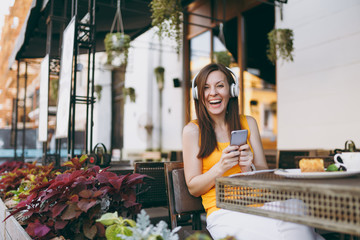 Pretty woman in outdoors street coffee shop cafe sitting at table, listen music in headphones, using mobile phone, relaxing in restaurant on free time. Mobile Office in summer. Lifestyle rest concept.
